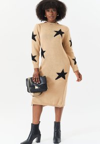 Beige knitted sweater dress with black star patterns, long sleeves, and ribbed neckline; paired with a black handbag and ankle boots.