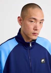 Casaco de pista Nike com base azul-marinho e painéis nos ombros azul-claro, gola alta, fecho de zip completo e um pequeno distintivo preto no peito.