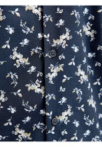 Blusa con motivo floreale in blu navy con tessuto testurizzato; presenta fiori crema e verdi, due bottoni neri e un design del colletto classico.