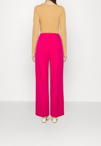 Femme portant un pantalon large rose vif, un haut beige à manches longues et des chaussures à talons larges couleur crème, se tenant devant un fond blanc uni.