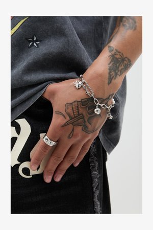 Silver bracelet with charms, including a bell and skull; worn on a hand displaying a wide silver ring. Gray shirt and distressed black jeans visible.
