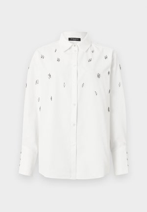 Chemise blanche à manches longues avec col et boutons, ornée de petites perles noires et argentées dispersées sur le devant et les manches.