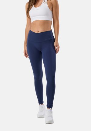Donna con reggiseno sportivo bianco, leggings blu navy a vita alta e sneakers bianche, in piedi contro uno sfondo chiaro e uniforme.