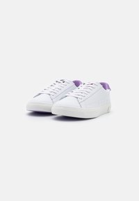 Un paio di sneaker bianche con lacci e fodera interna viola su uno sfondo bianco, vista angolata frontale.
