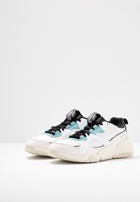 Sneaker bianchi e azzurro chiaro con accenti neri, realizzati in materiali in rete e pelle, con sovrapposizioni testurizzate e un sistema di allacciatura audace.