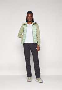 Casaco puffer verde claro com uma textura suave, combinado com uma camisa branca e calças escuras justas. Ténis refletivos com detalhes roxos.