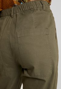 Kaki byxor med elastisk midja, med en bakficka och en slät yta. Sömdetaljer runt fickans kanter är synliga.