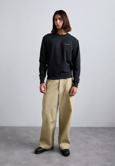 Black sweatshirt with a crew neck and logo, paired with beige wide-leg trousers and black slip-on shoes. Smooth fabric, relaxed fit.