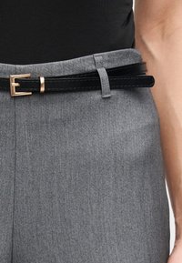 Ceinture en cuir noire avec boucle dorée portée avec un pantalon gris taille haute et un haut noir rentré, montrant un gros plan de la taille et du bras.