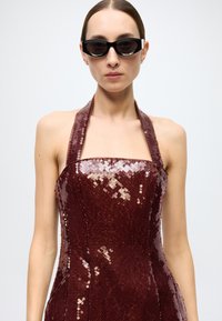 Burgundy sequin dress with a fitted bodice, halter neck design, and shiny texture. Black cat-eye sunglasses complete the look.