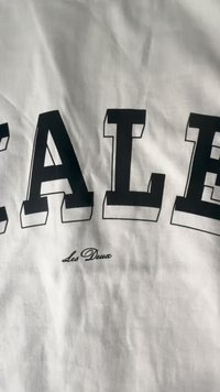 Tecido branco com letras em negrito, pretas e em estilo de bloco a soletrar "SALE", com uma textura ligeiramente elevada e um logotipo em script delicado abaixo.
