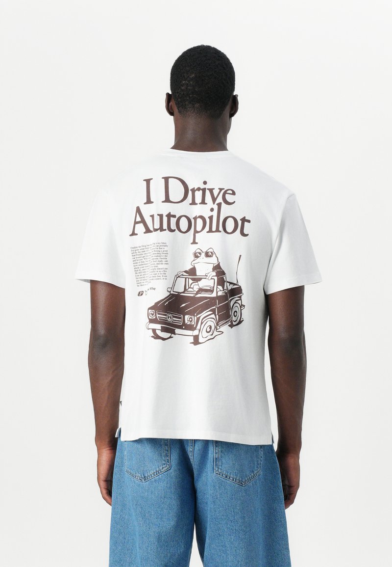 Weißes Baumwoll-T-Shirt mit einem großen braunen Grafikmotiv auf dem Rücken, das einen Frosch zeigt, der ein schwarzes Auto fährt, mit Text über und unter dem Bild.