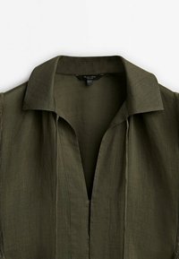 Olivenfarvet, kortærmet skjorte med fold-down krave og åben front uden knapper. Fremstillet af letvægtsstof med en glat tekstur.