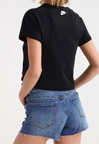 Person wearing black cropped Nike t-shirt and high-waisted blue denim shorts, standing with hands in pockets against white background.