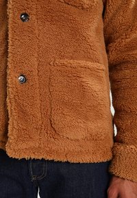 Veste en polaire marron avec une finition douce et texturée, présentant des fermetures à boutons noirs et un contour de poche avant.