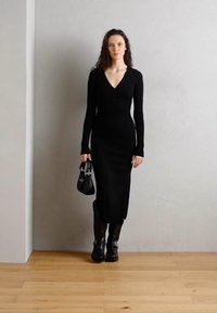 Black knitted dress with long sleeves and a deep V-neck, featuring button accents, side slits, paired with black knee-high boots and a handbag.