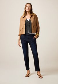 Chaqueta corta de ante color marrón, cárdigan gris, top blanco sin mangas, pantalones oscuros de mezclilla y chanclas negras; diseño simple con mínima ferretería.