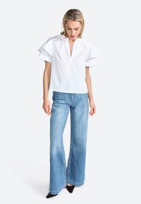 Blusa bianca a maniche corte con polsini arricciati e scollatura a V, abbinata a jeans a gamba larga blu chiaro e scarpe nere a punta.