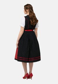 Zwarte jurk met bloemenpatroon, rode accenten en aansluitende corsage. Witte blouse met gerimpelde mouwen. Rode schoenen.