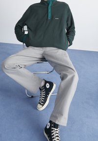 Mørkegrøn halvmole pullover i fleece med teal detaljer, grå bukser og sorte højhælede sneakers mod en blå baggrund.