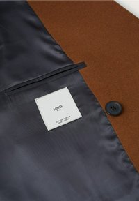 Intérieur d'une veste en mélange de laine marron avec doublure en satin foncé, bouton et étiquette d'entretien. Comprend une seule poche et une texture lisse.