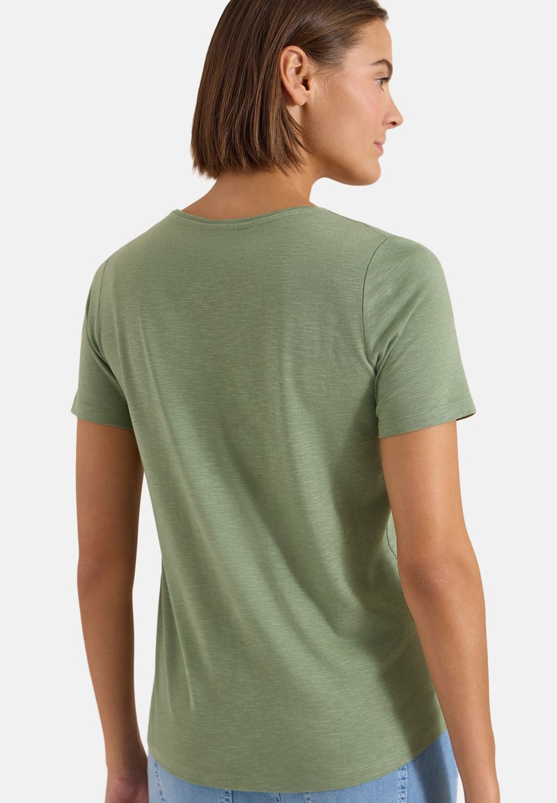 Femme aux cheveux bruns courts portant un t-shirt vert uni à manches courtes, vue de dos et légèrement tournée sur le côté.