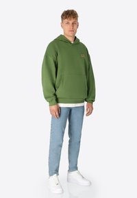 Grüne Kapuzenjacke mit einer Vordertasche und einem kleinen Logo, getragen über einem weißen Shirt, kombiniert mit hellblauen Jeans und weißen Turnschuhen.