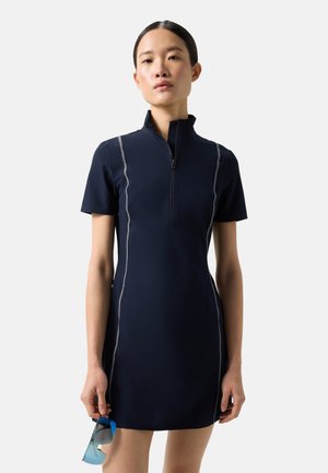 Kvinde iført en tætsiddende navy kortærmet kjole med hvide sidelæg, holder blåtonede solbriller i højre hånd mod hvid baggrund.