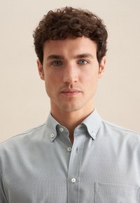 Camicia azzurra chiara con tessuto testurizzato, colletto corto e taschino sul petto. Presenta bottoni bianchi e dettagli di cucitura.