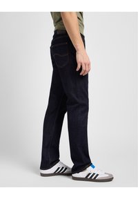 Jeans de mezclilla azul oscuro con un corte recto, que presentan detalles de costura y un diseño de bolsillo trasero, combinados con zapatillas blancas y grises.
