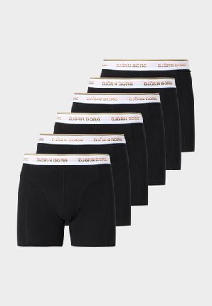 Jeune homme noir debout portant un boxer noir avec une ceinture blanche et orange, regardant vers sa gauche sur un fond clair uni.