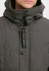 Chaqueta aislante gris con un alto cuello, capucha ajustable con cordón, cierre de cremallera, botones a presión y bolsillos frontales con acentos cosidos.