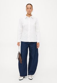 Hvit langermet skjorte med subtil tekstur, kombinert med brede mørkeblå denimbukser. Modellen holder en liten håndveske med franjene.