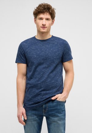 Young man with curly hair wearing a blue speckled t-shirt and jeans, standing with one hand in pocket against a plain background.