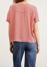 Kvinna som bär en röd och vit vertikalt randig kortärmad skjorta och blå denimjeans, visad bakifrån mot en enkel bakgrund.