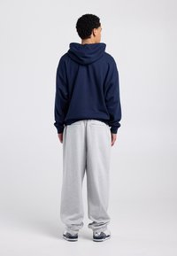 Sweat à capuche marine avec une grande poche arrière ; associé à un pantalon de survêtement gris clair. Tissu lisse, coupe décontractée et poignets élastiques aux chevilles.