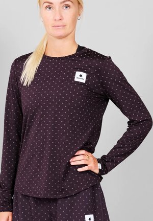 Frau mit blondem Pferdeschwanz, die ein schwarzes Langarm-Polka-Dot-Shirt und passende Hosen mit dem "SAYSKY"-Logo auf Brust und Oberschenkel trägt.