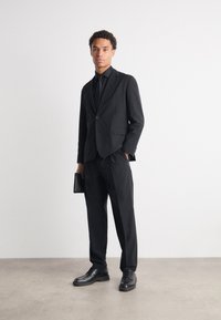 Black tailored suit with a single button, matching tie, and polished black shoes. Holding a small black clutch, standing on a neutral background.