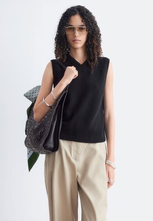 Personne aux cheveux bouclés portant des lunettes de soleil, un haut noir sans manches, un pantalon beige, et tenant un grand sac noir texturé avec un foulard à motifs.