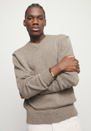 Joven con cabello trenzado, vestido con un suéter de punto beige y pantalones blancos, con los brazos cruzados, mirando directamente al frente sobre un fondo liso.