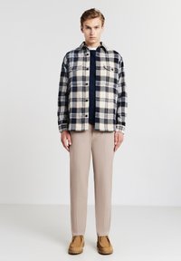 Camicia di flanella a plaid in navy, crema e marrone, con chiusura a bottoni; camicia navy sotto. Pantaloni beige e scarpe in suede marroni completano l'outfit.