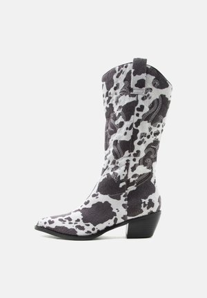 Tehénmintás bokáig érő cowboy csizma hegyes orral, blokksarokkal, díszes fehér varrással és húzófüllel világos háttéren.