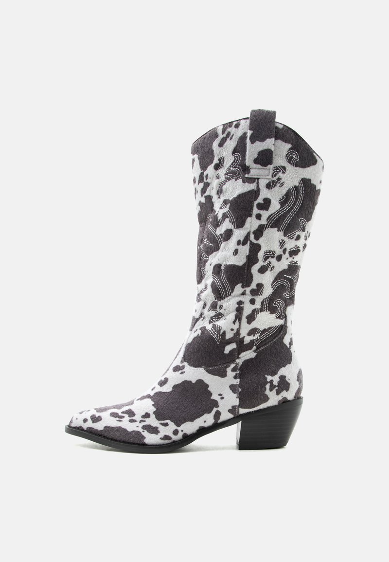 Botte de cowboy mi-mollet imprimé vache, avec bout pointu, talon bloc, couture blanche décorative et languette d'extraction sur fond clair.