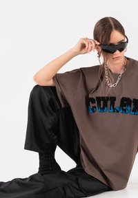 Brązowy, oversize t-shirt z napisem "CUL" w czarnym i niebieskim projektu płomieni. Czarnie luźne spodnie, warstwowy naszyjnik ze srebrnych łańcuchów oraz czarne okulary przeciwsłoneczne.