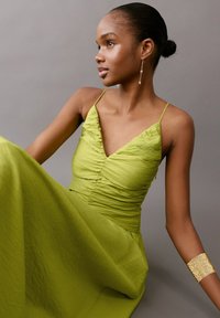 Ženska s temno poltjo in urejeno figo, oblečena v brezrokavno limetino zelene barve obleko, z visečimi uhani in široko zlato zapestnico, sedi pred sivim ozadjem.