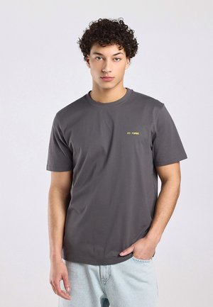 Junger Mann mit lockigem Haar, der ein dunkelgraues T-Shirt mit gelbem Text "NYC - FLOWROS" und hellblaue Jeans trägt, steht mit einer Hand in der Tasche.