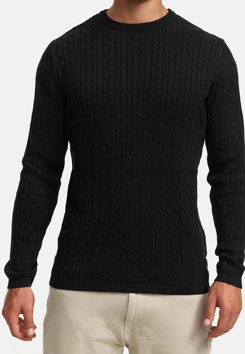 Homme portant un pull noir à manches longues en maille torsadée avec poignets et ourlet côtelés, associé à un pantalon de couleur claire.