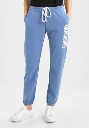Osoba nosząca niebieskie spodnie dresowe z białym sznurkiem i logo Abercrombie na udzie, w połączeniu z białymi trampkami i czarnym topem.