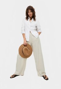 Biała koszula na guziki, luźne jasnobeżowe spodnie, czarne klapki oraz jasnobrązowy kapelusz ze słomy. Model stoi w swobodnej pozie na jednolitym tle.