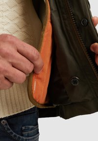 Olivově zelená bunda s oranžovou podšívkou, zobrazující detail knoflíku a kontrastní lemování. Tkanina působí hladce se strukturovaným střihem.